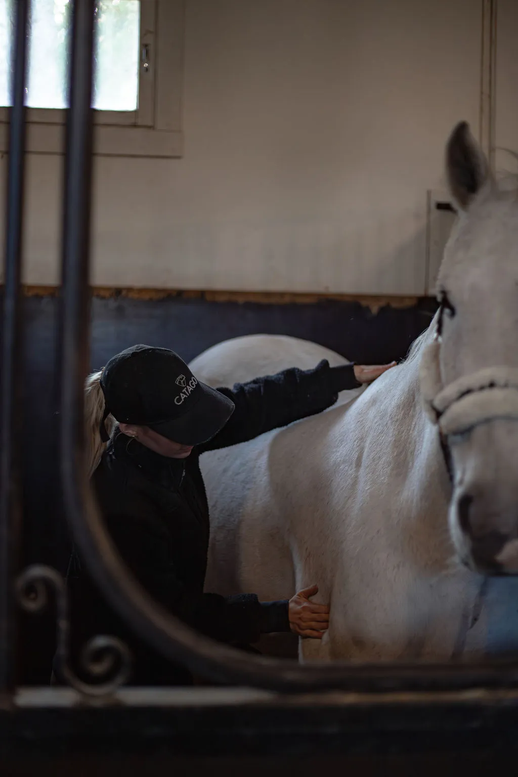 Tallissa oleva valkoinen hevonen, jota hoitaja silittää, näkymä tallin portin läpi.